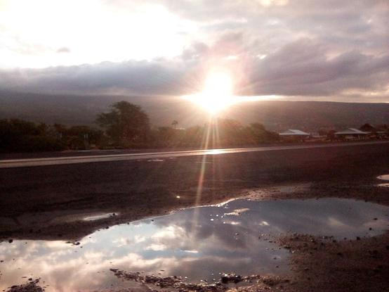 A Photo of the Sun rising over the mountains, shining through the clouds. A reflection of the clouds is seen in a puddle.

Photo taken in Kailua-Kona, Hawai'i